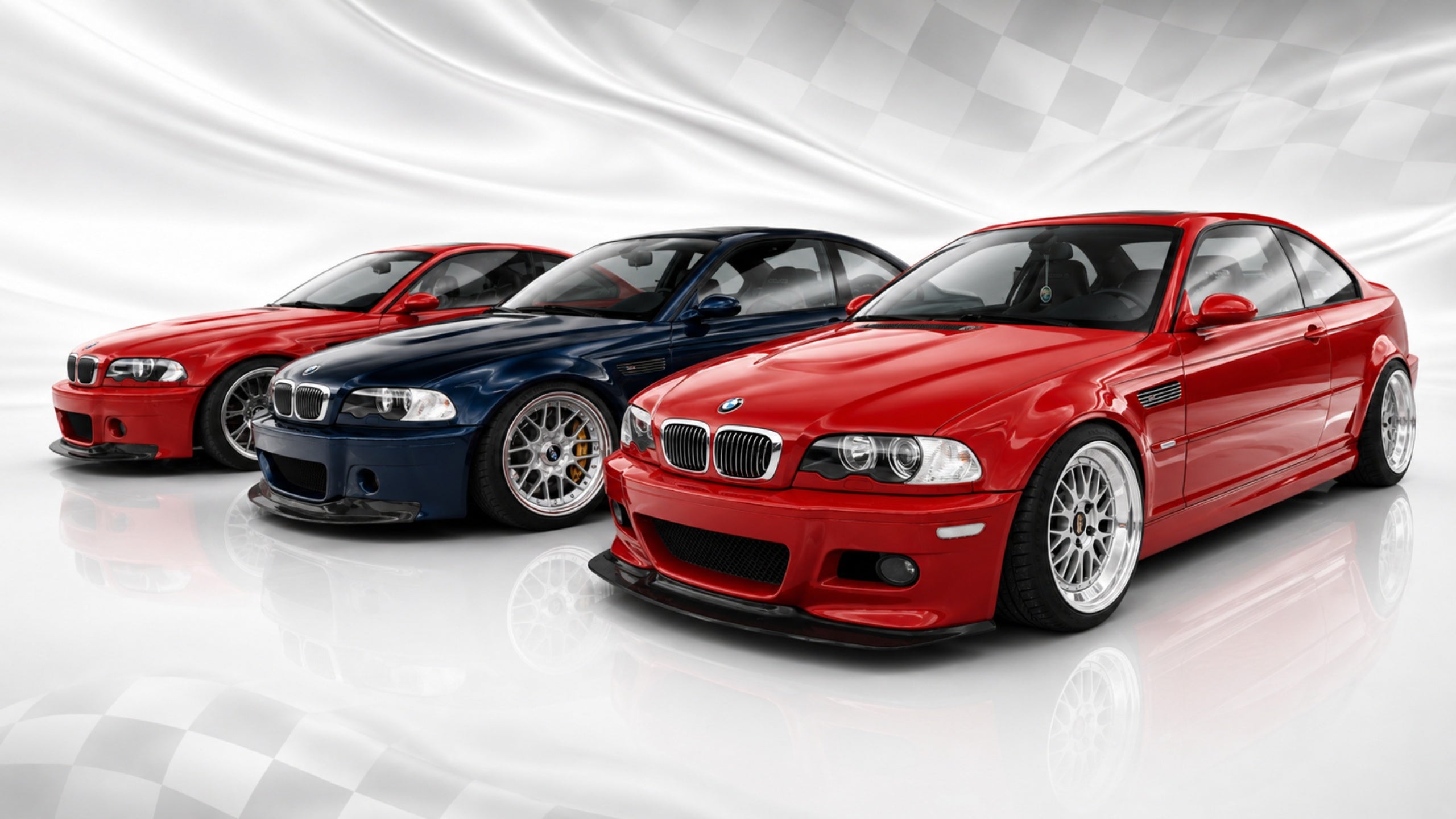 Three BMW M3 coupes — two red and one dark blue — lined up against a checkered racing flag backdrop. Join AutomotiveMust Rewards to earn points on every order.